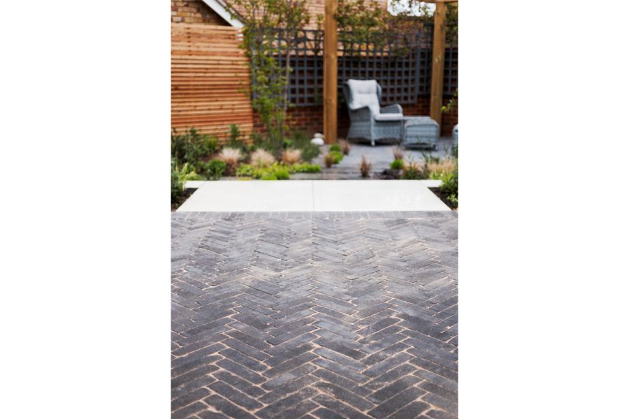 Wide path of Lucca clay pavers laid herringbone leads to pale paving with staggered path and patio with furniture beyond.