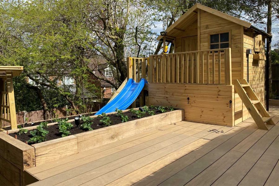 Garden decking of Warm Teak Brushed Designboard with large chalet-style playhouse and raised bed edged with wooden sleepers.