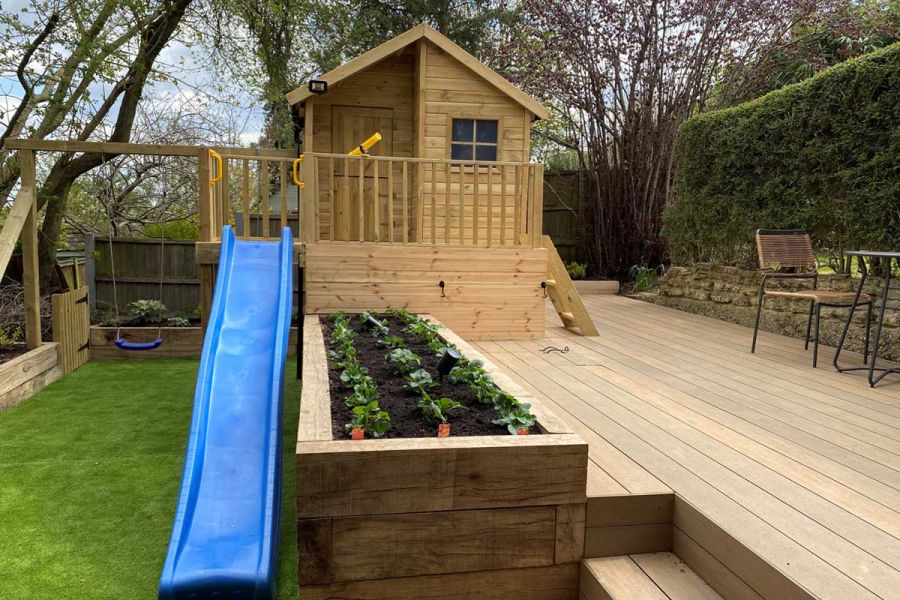 Wooden playhouse on raised terrace of Warm Teak brushed DesignBoard, next to blue slide, swing, raised beds and small lawn.