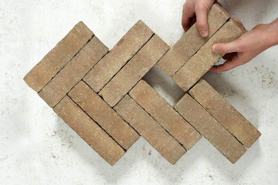 2 hands add 2 bricks to 10 Westminster Clay Pavers in double herringbone pattern on grey background. Next-day delivery available.