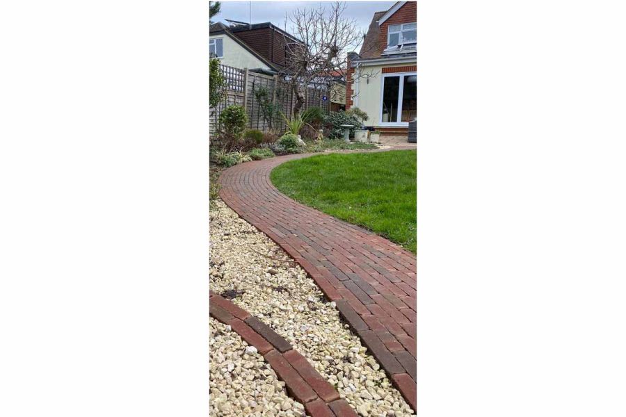Curved garden path in Winton brick pavers leads from patio doors of suburban house between lawn, beds and pale gravel.