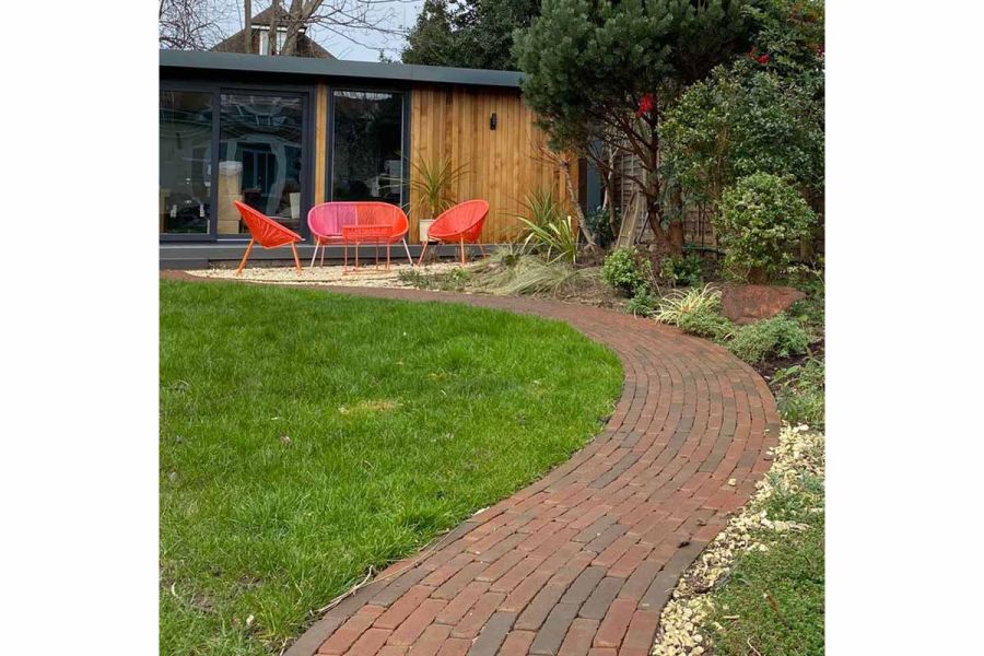 Curved path of Winton brick pavers leads between lawn and bed to garden studio with outdoor furniture. Design by Landscape Artisan.