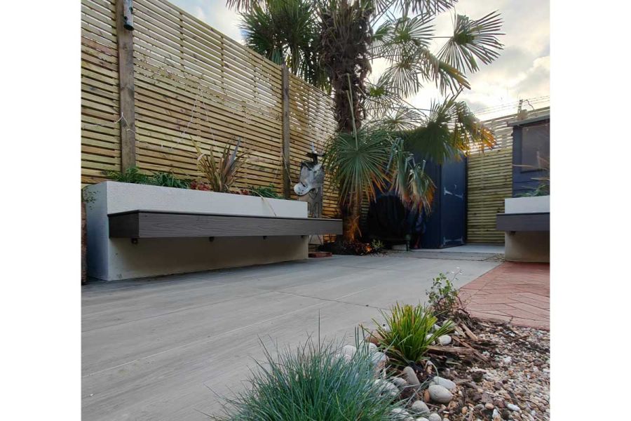 Nuage Porcelain Paving edged on 2 sides with tall slatted fencing, with dry planting and palm trees. Designed by Floral and Hardy.