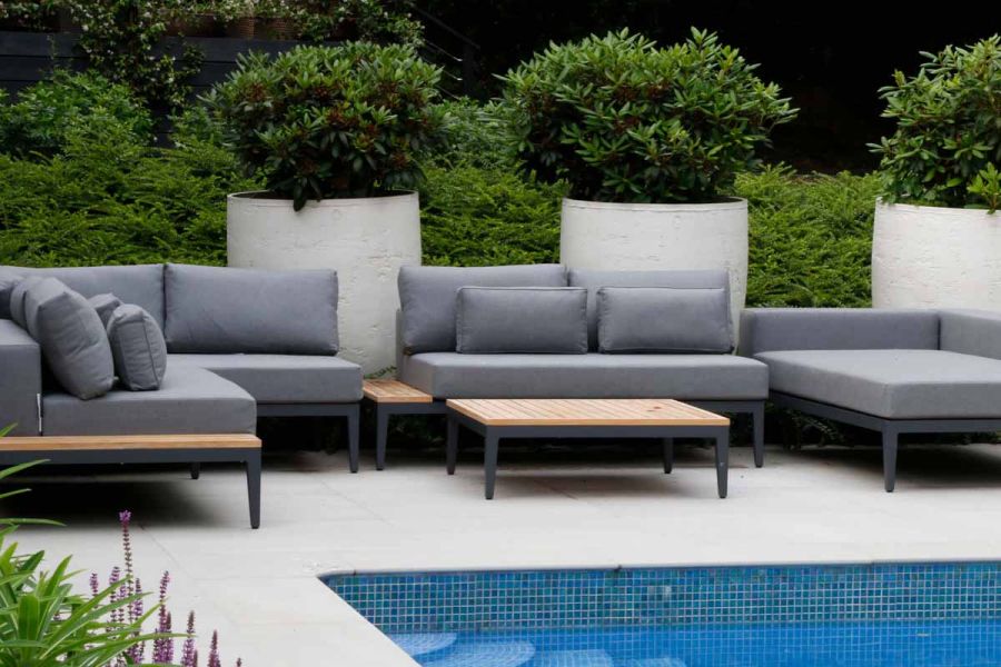Modern grey cushioned outdoor furniture sits next to pool clad with Yard 1200x1200 Porcelain Paving with large trees in the background.