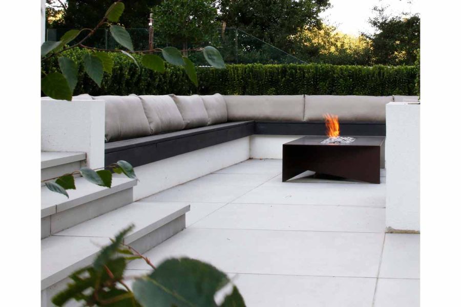 Low view of Yard 1200x1200 Porcelain Paving with built in bench with white cushions, surrounding a cube firepit.