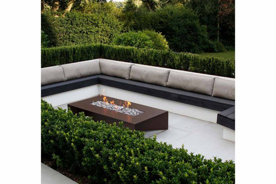 Fire roars in corten clad firepit with pebbles. Green hedges surround a built in bench with cushions on top of yard 1200x1200 porcelain paving.