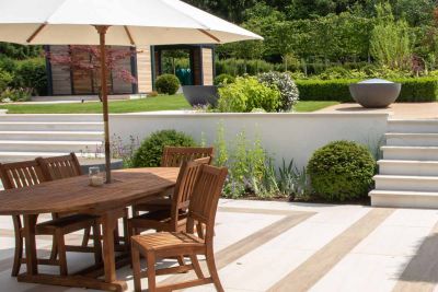 Wooden table and chair set with erected umbrella has glorious views of raised patio showing off Faro Porcelain Wall Copings.***Designed by Caroline Davy Studio, www.instagram.com/carolinedavystudio | Built by PC Landscapes Ltd, www.pclandscapes.co.uk
