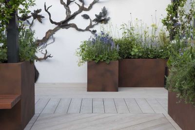 In front of white wall with metal sculpture attached, corten steel planters sit on Limed Oak Millboard decking laid in 3 directions.***Designed by Rhiannon Williams | Built by Garden Club London Ltd, www.gardenclublondon.co.uk
