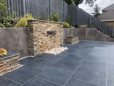 Charcoal porcelain paving patio in front of a stone clad water feature and large retaining wall clad with large format porcelain cladding.***Manor Prestige Landscapes, www.manor-prestige-landscapes-ltd.business.site