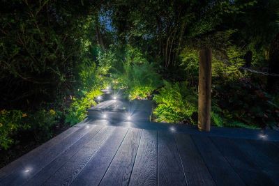 Night view of woodland borders on either side of staggered sections of Burnt Cedar Millboard decking with inset light spots.***Monarch Lawn & Landscape, www.monarchlandl.co.uk