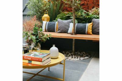 Round yellow coffee table on Slab Coke Porcelain outdoor tiles UK. Behind, small tree grows through hole in wooden bench seat.***Georgia Lindsay,  www.georgialindsaygardendesign.com