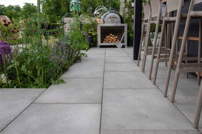 Near-ground view trade stand laid with Venetian Grey porcelain vitrified paving, with planted bed, legs of bar set and pizza oven.***Garden House Design, www.gardenhousedesign.co.uk