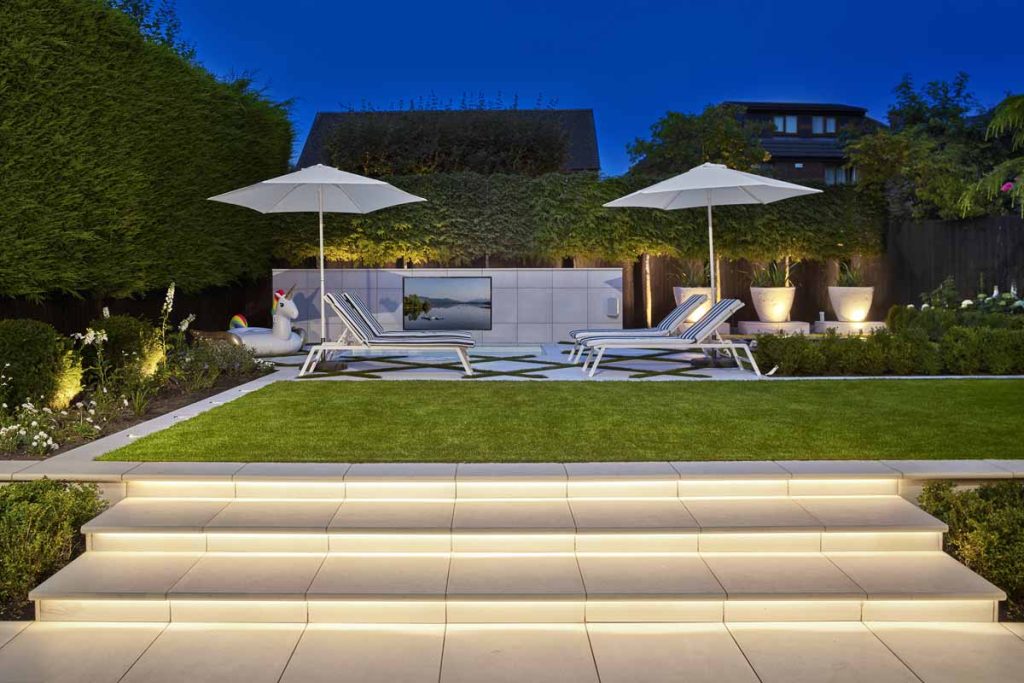 Wide, underlit steps of Comblanchien porcelain rise to rectilinear lawn with 2 parasols and sunloungers.