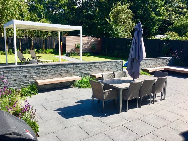 Large patio in Trendy Black porcelain square patio tiles laid running bond, edged with stone clad wall and lawn beyond.