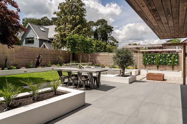 Sidewalk large format porcealin paving by white raised bed, lawn and pergola-covered area