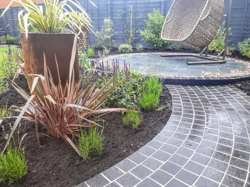 curved path of crisply pointed setts leads to round patio of Slab Coke porcelain paving with egg chair. 