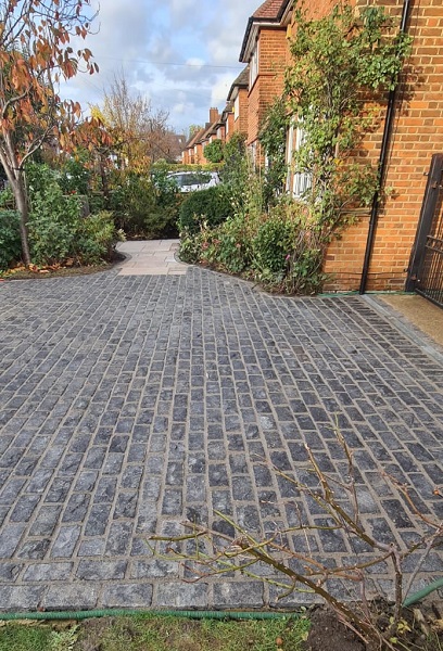 View across wide driveway of black granite setts to front garden of brick house.