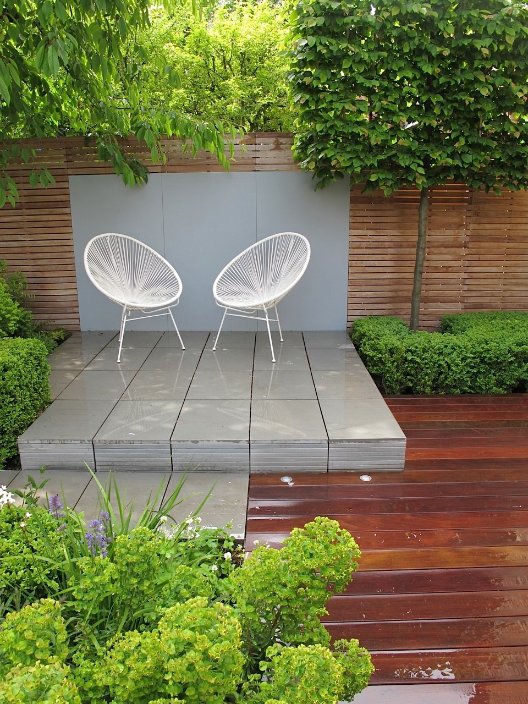 Raised Contemporary Grey sawn sandstone patio with 2 white chairs with clipped hedging on either side.