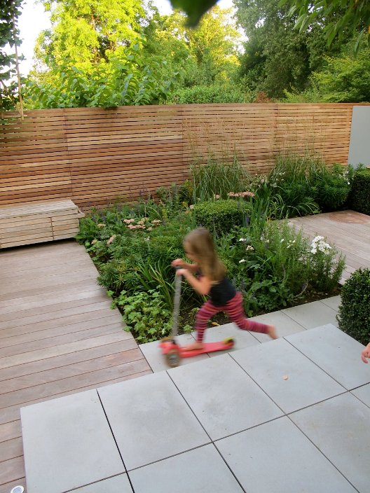 Girl on scooter rushes past Contemporary Grey sawn sandstone patio to decked area.