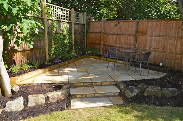 2 steps up to triangular Mint Indian sandstone patio in fenced corner. Fig tree on left. 