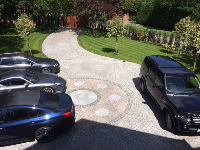 Long curved driveway leads to turning circle with pattern of mixed paving setts and parked cars.