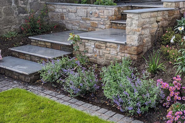 Mowing strip of 2 courses of natural Black Granite paving setts separates lawn from plant border at base of flight of steps.