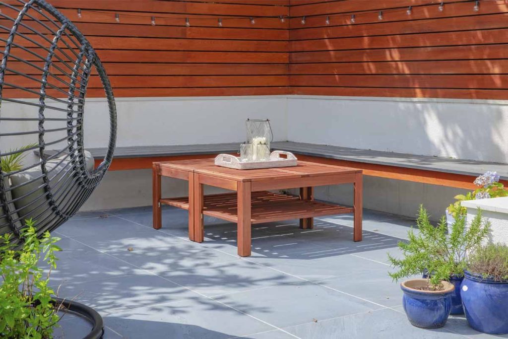 Kirkby Porcelain paving for patios laid in corner of garden with slatted fencing and wooden table. Design by Mustard Seed.