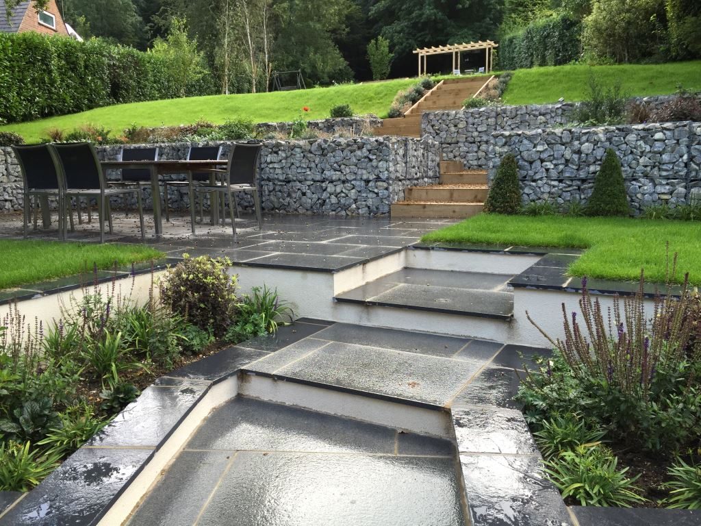 Blue grey granite steps and patio below gabion-retained rising lawn, with black limestone coping on raised beds.