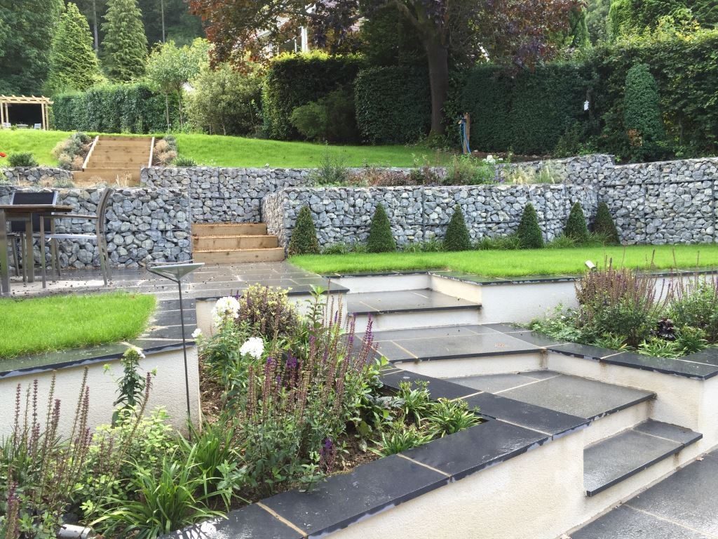 Terraced sloping garden with blue-grey granite steps and white rendered walls leading up to retaining wall of gabions. 