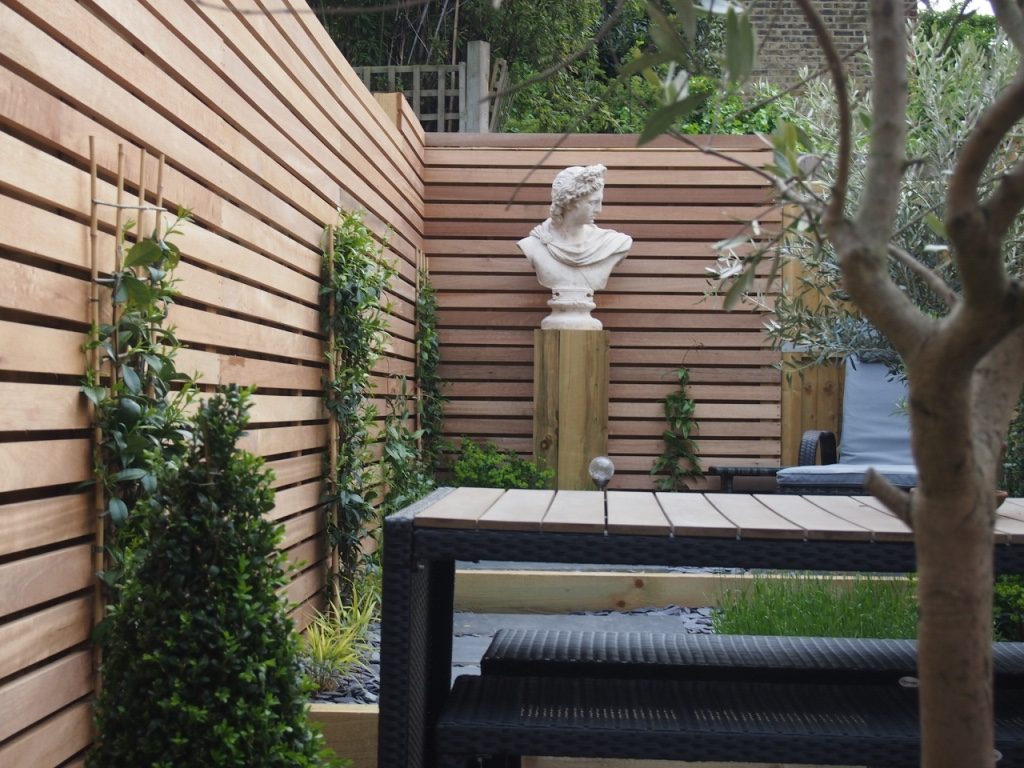 Bust on plinth in corner of fenced garden with wood-topped table and slate paving.