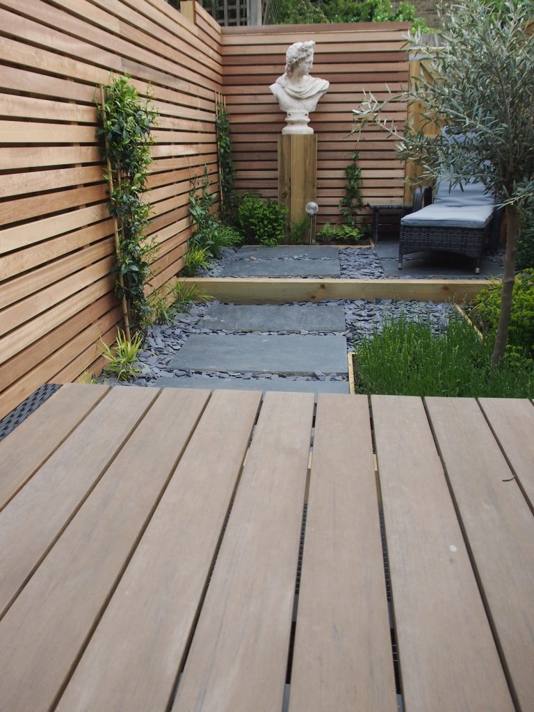 Black slate paving and chipping seen beyond wooden tabletop, with step up to next level of small sloping garden.