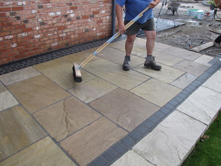 Man in shorts removing residual GftK brush-in patio grout with a brush from sandstone paving.