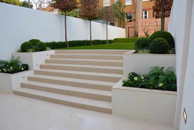 Oblique view of 8 Warm Beige porcelain steps rising between raised beds to artificial lawn between rendered garden walls. 