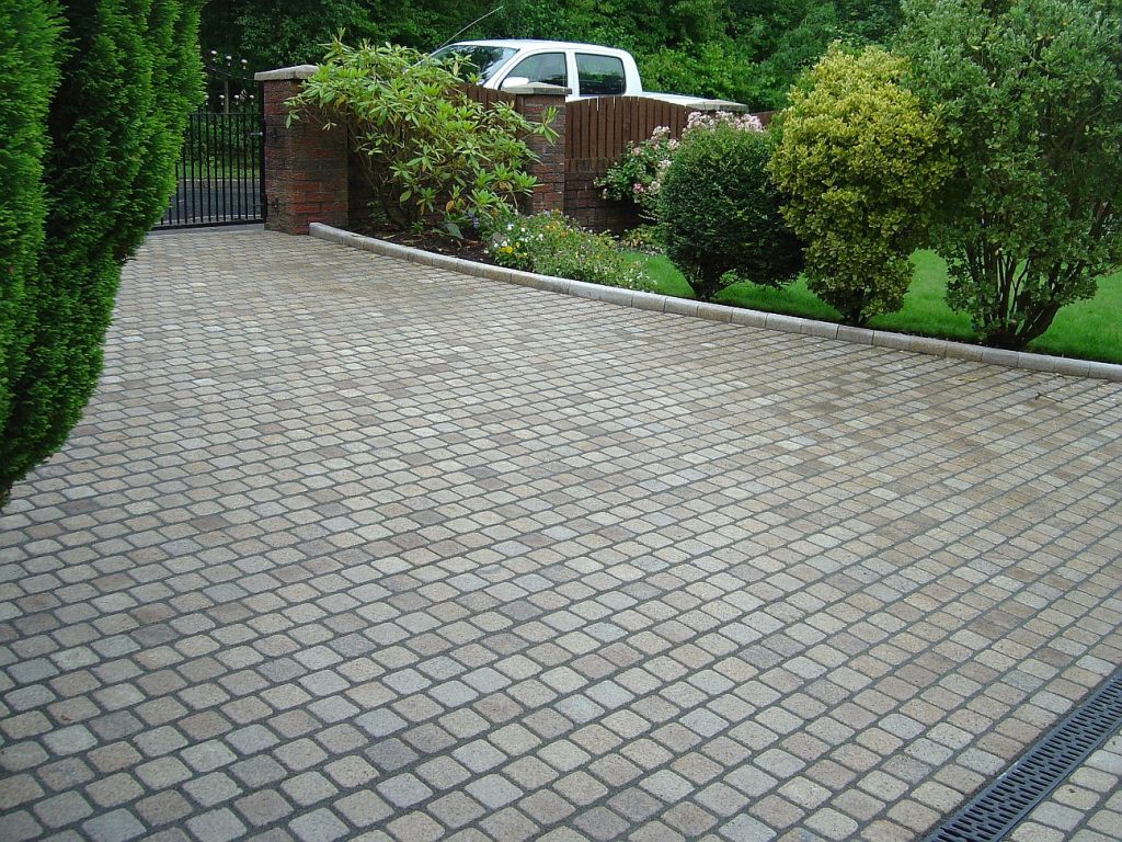 Driveway of granite setts, bushes on far side, metal gate onto road, with brush-in patio grout.