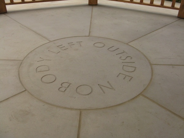Round Buff Yorkstone paving stone, engraved with 'Nobody Left Outside', in middle of radial-cut slabs.