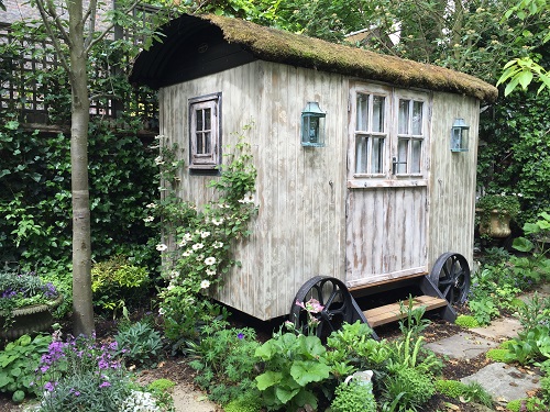 A shepherd hutwith distress paint  sits on reclaimed paving with naturalistic planting. 