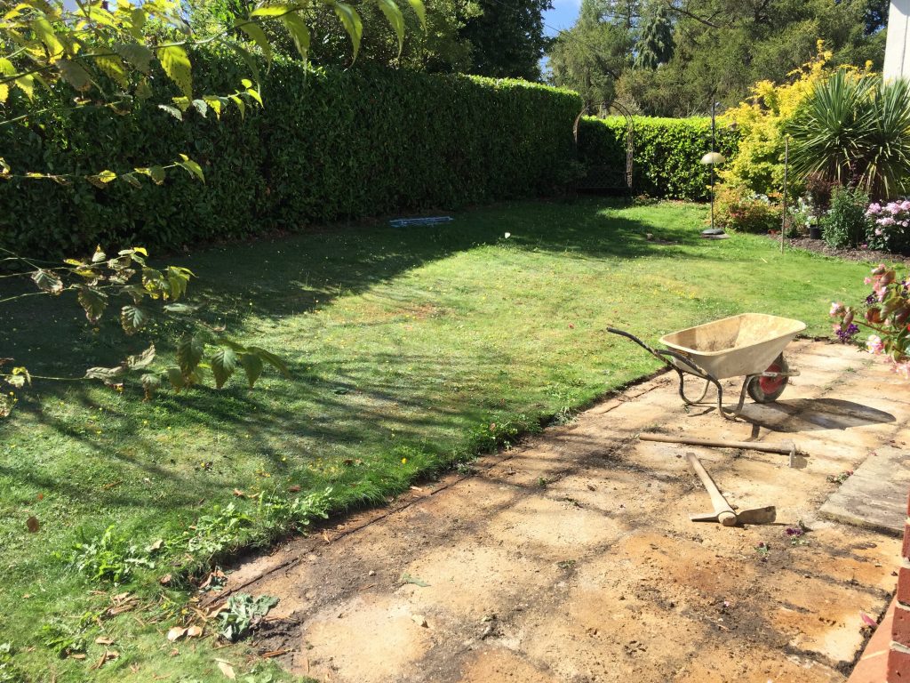 Wheelbarrow sits on rectangle of bedding mortar where old patio has been removed, next to lawn and hedge.