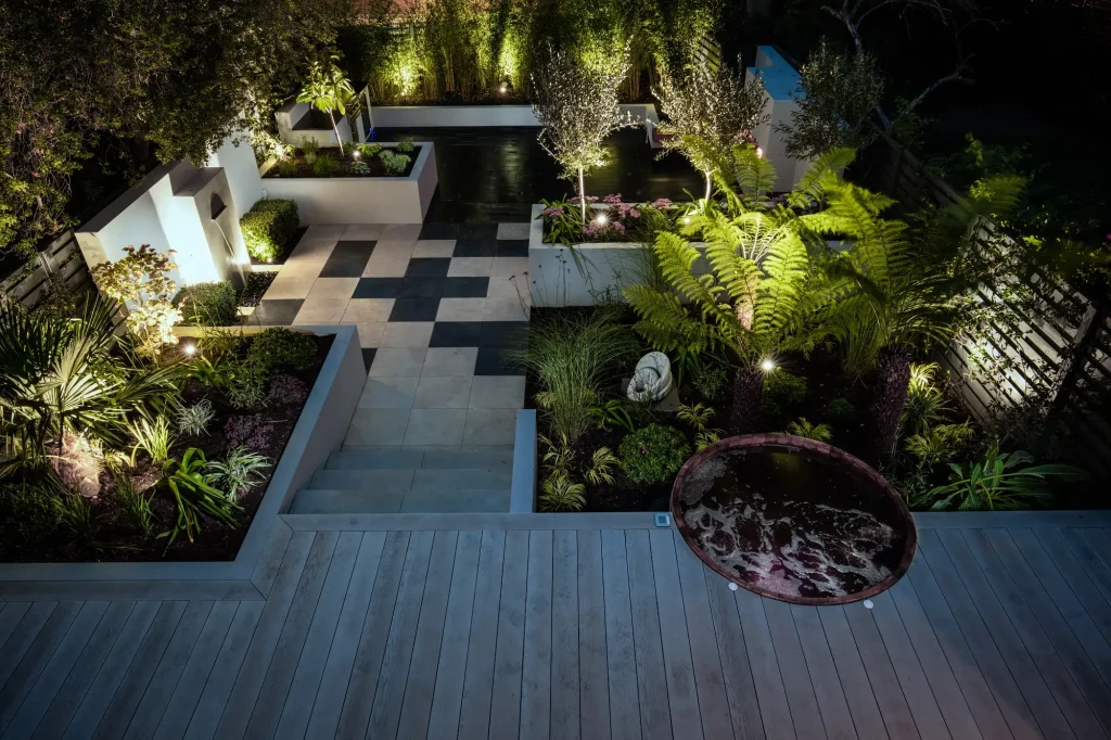 Roof terrace with steps down from decking to mix and match patio slabs in black and white. Raised planters and uplit trees.