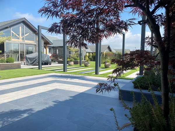Large square paved area in charcoal porcelain paving with light grey stripes next to path with modern pergola. Lawn beyond.