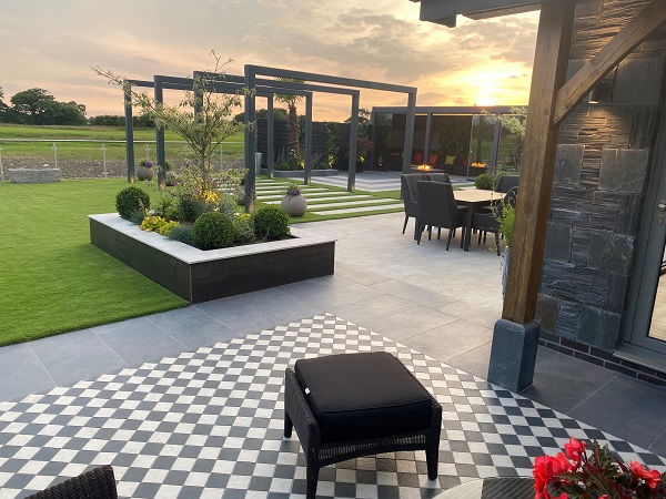 Stool sits on Charcoal and Light Grey porcelain setts in chequerboard pattern, with large patio and pergola covered path behind. 