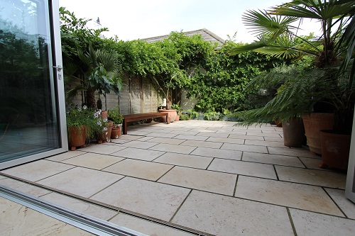 View through open bifold doors, over stainless steel slot drain, to small garden totally paved with Jura Beige Limestone.