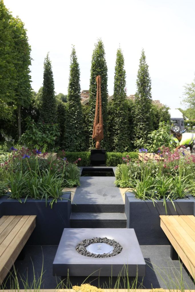 Statue of man hangs upsidedown over paving with planted borders on either side