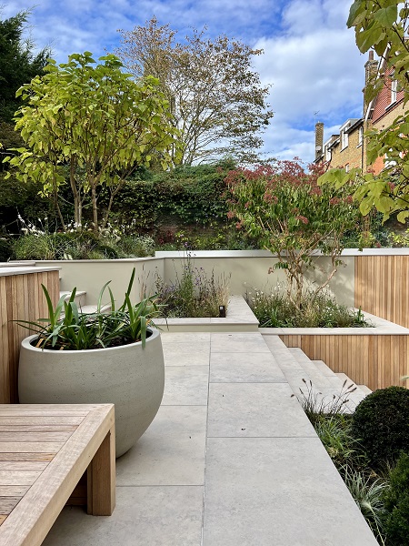 Steps rise to narrow terrace paved with Jura Grey Italian outdoor tiles. Wooden bench and large white pot to left.