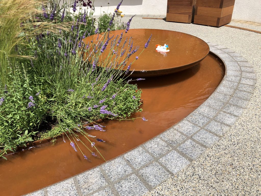 Corten Steel shallow bowl water feature set within pond edged with Silver-Grey Granite Setts. RHS Hampton Court 2019