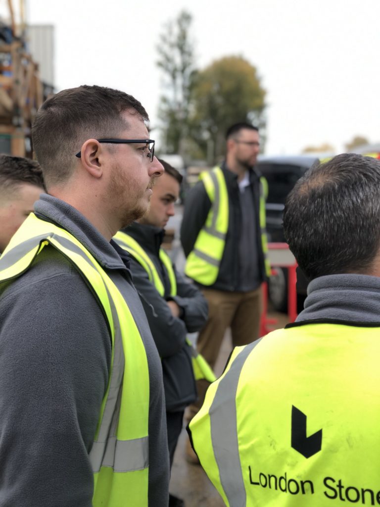Row of men in hi-vis vests in the open air, all looking at something out of the frame.