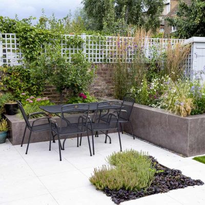 Raised beds faced with Vulcano Ceniza Designclad porcelain wall cladding next to metal dining set on white paving with inset bed.