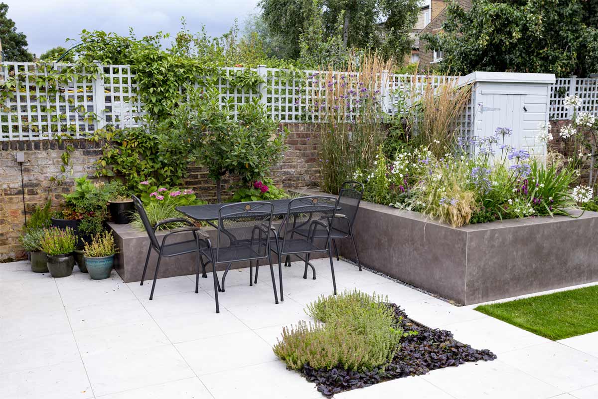 Raised beds faced with Vulcano Ceniza Designclad porcelain wall cladding next to metal dining set on white paving with inset bed.