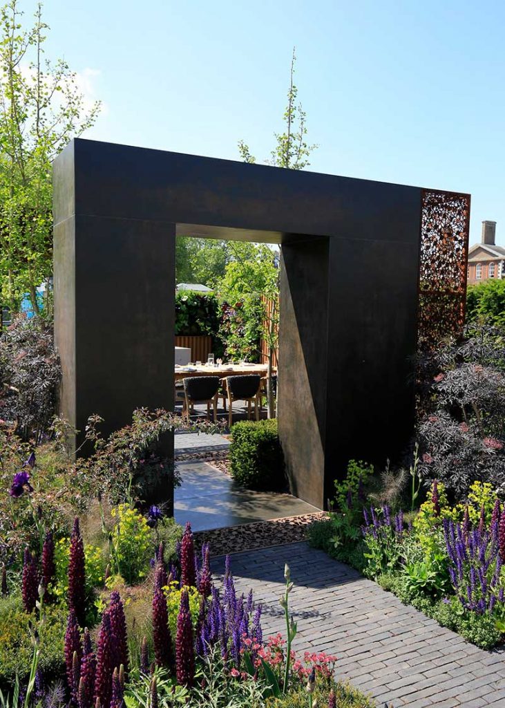 Clay paver path leads to dining area through arch faced with Steel Dark DesignClad in RHS Chelsea 2018 garden by Tony Woods.