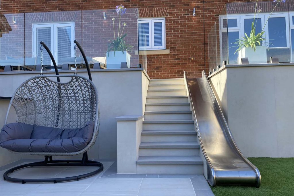 10 Steps with polished steel children's slide descend between flanking walls from patio by house to lawn and paving.