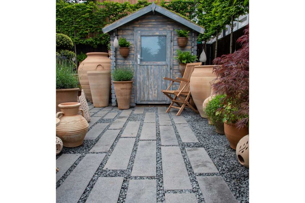 Dark grey granite planks point to shed with terracotta pots, with wide gravel joints.